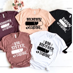 Puede incluir: Cinco camisetas con un tema de anuncio de embarazo. Las camisetas son de diferentes colores y presentan el texto "Mommy Loading", "Daddy Loading", "Grandma Loading", "Big Sister Loading" y "Big Brother Loading".