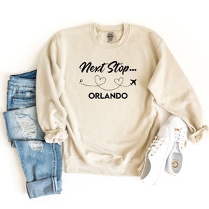 May include: A beige sweatshirt with the text "Next Stop... ORLANDO" in black. The text is accompanied by a graphic of a plane flying over two hearts.