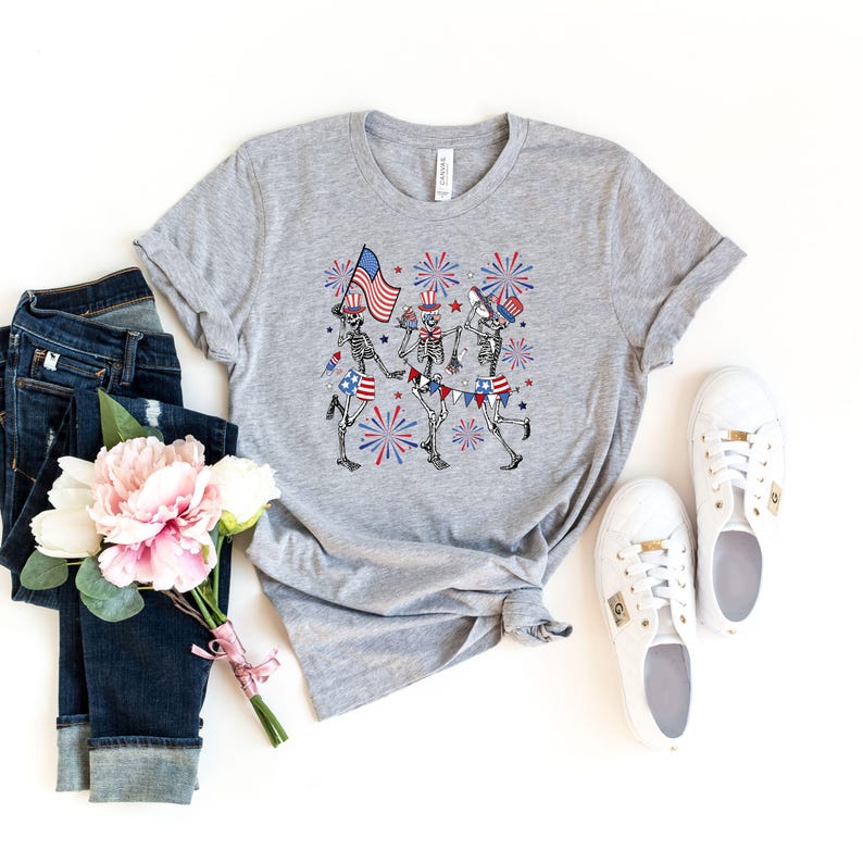 May include: A heather gray t-shirt featuring a festive design of skeleton figures celebrating with American flags and fireworks. The design incorporates red, white, and blue colors, perfect for patriotic events. The shirt is paired with blue jeans and white sneakers.