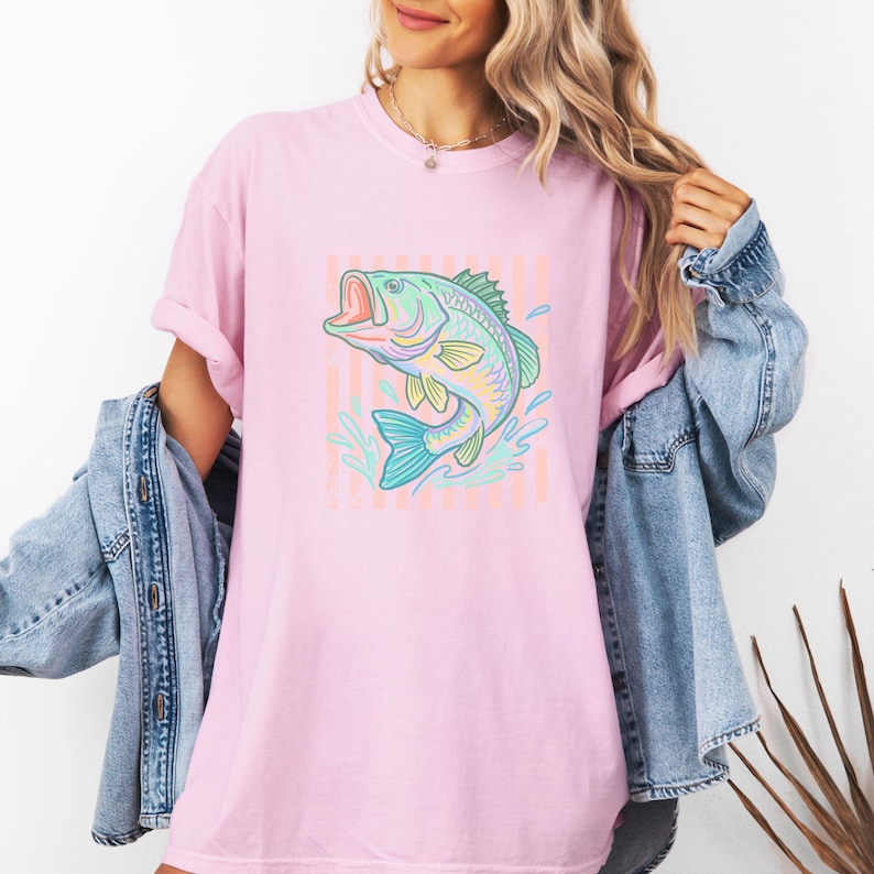 Pu&ograve; includere: T-shirt rosa chiaro con un'illustrazione colorata di un pesce che salta. Il pesce &egrave; in tonalit&agrave; di blu, verde e giallo, con spruzzi d'acqua. Lo sfondo presenta strisce verticali rosa e pesca.