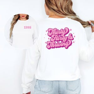 May include: White long-sleeve sweatshirt with the text "Thirty, Flirty & Thriving" in pink and purple on the back. The front of the sweatshirt has the number "1995" in pink. The sweatshirt is a casual style.