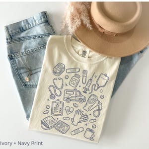 May include: An ivory-colored t-shirt with a navy print featuring medical-themed illustrations, including a stethoscope, ambulance, and medicine bottles. The shirt is paired with light blue jeans and a tan hat. The text "Ivory + Navy Print" is at the bottom.