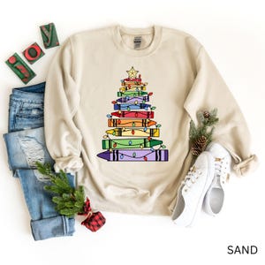 May include: A sand-colored sweatshirt features a Christmas tree design made of colorful crayons. The crayons are stacked to form the tree shape, adorned with string lights and topped with a gold star. The word "JOY" is visible in the upper left corner.