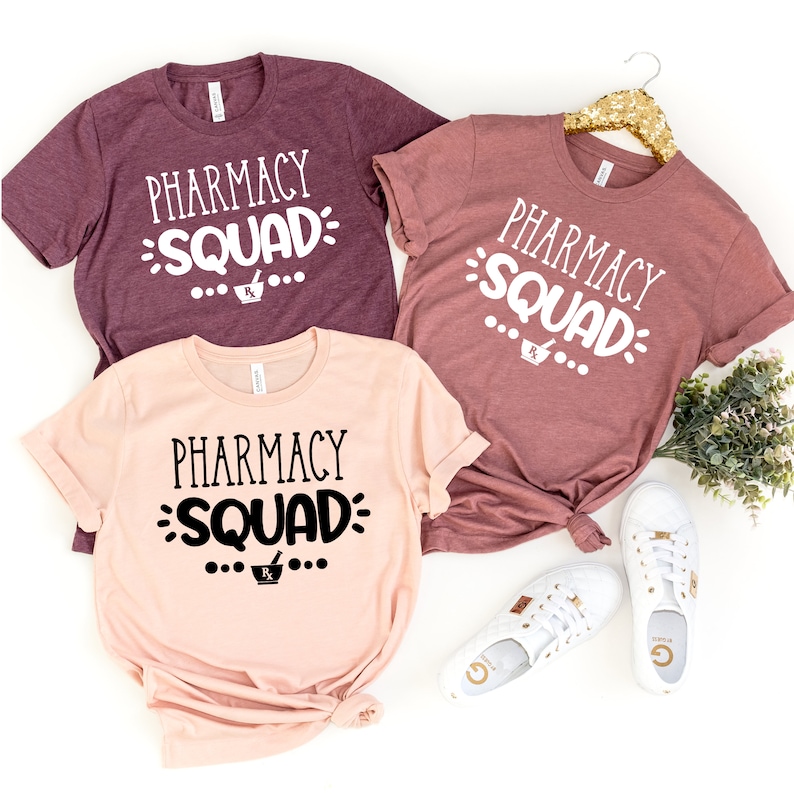May include: Three t-shirts with the text "Pharmacy Squad" in black and white lettering. The shirts are in different colors: maroon, pink, and a darker shade of pink. The shirts are laid out on a white background with white sneakers.