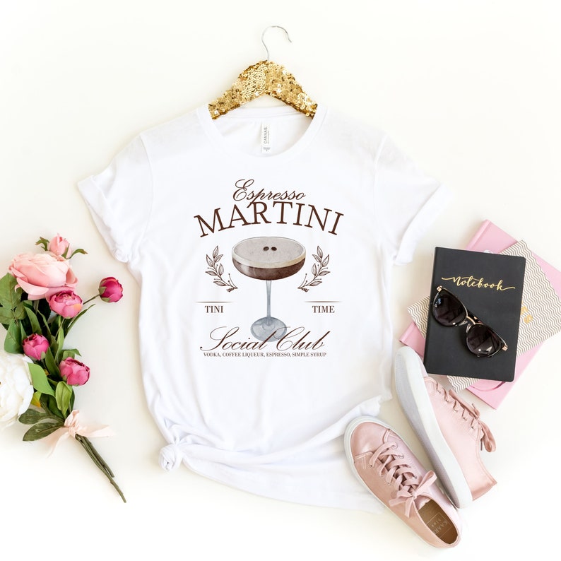 May include: White t-shirt with a brown graphic of an espresso martini cocktail. The graphic includes the text "Espresso Martini", "Tini Time", "Social Club", and "Vodka, Coffee Liqueur, Espresso, Simple Syrup".