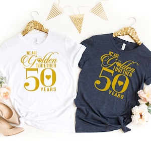 Pode incluir: Duas camisetas com um gr&aacute;fico dourado que diz "We are Golden Together 50 Years" com um diamante e um cora&ccedil;&atilde;o. As camisetas s&atilde;o brancas e azul escuro.