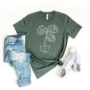 May include: Green t-shirt with a white line drawing of a potted Monstera plant. The shirt is styled with light blue ripped jeans and white sneakers. The shirt is knotted at the waist.