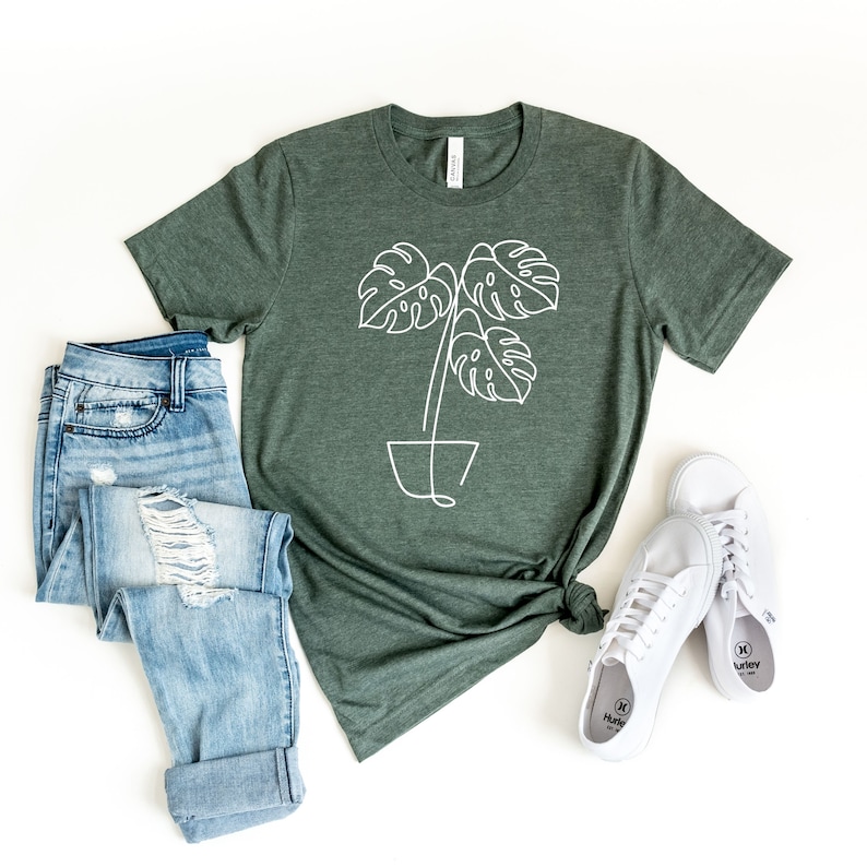 May include: Green t-shirt with a white line drawing of a potted Monstera plant. The shirt is styled with light blue ripped jeans and white sneakers. The shirt is knotted at the waist.