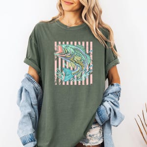 Pu&ograve; includere: T-shirt verde oliva con una grafica colorata di un pesce che salta con la bocca aperta. Il pesce &egrave; posto su uno sfondo a strisce rosa e bianche. La maglietta ha maniche arrotolate e una vestibilit&agrave; ampia.