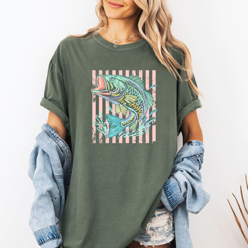 Pu&ograve; includere: T-shirt verde oliva con una grafica colorata di un pesce che salta con la bocca aperta. Il pesce &egrave; posto su uno sfondo a strisce rosa e bianche. La maglietta ha maniche arrotolate e una vestibilit&agrave; ampia.