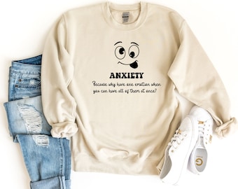 May include: A beige sweatshirt with a black graphic of a smiley face with a tongue sticking out and the text "ANXIETY Because why have one emotion when you can have all of them at once?"