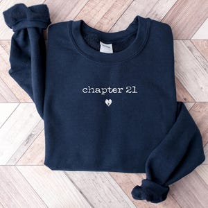 May include: Navy blue sweatshirt with the text "chapter 21" in a white, delicate font, and a small heart beneath it. The sweatshirt has a crew neck and ribbed cuffs. The garment is folded on a light brown wooden surface.