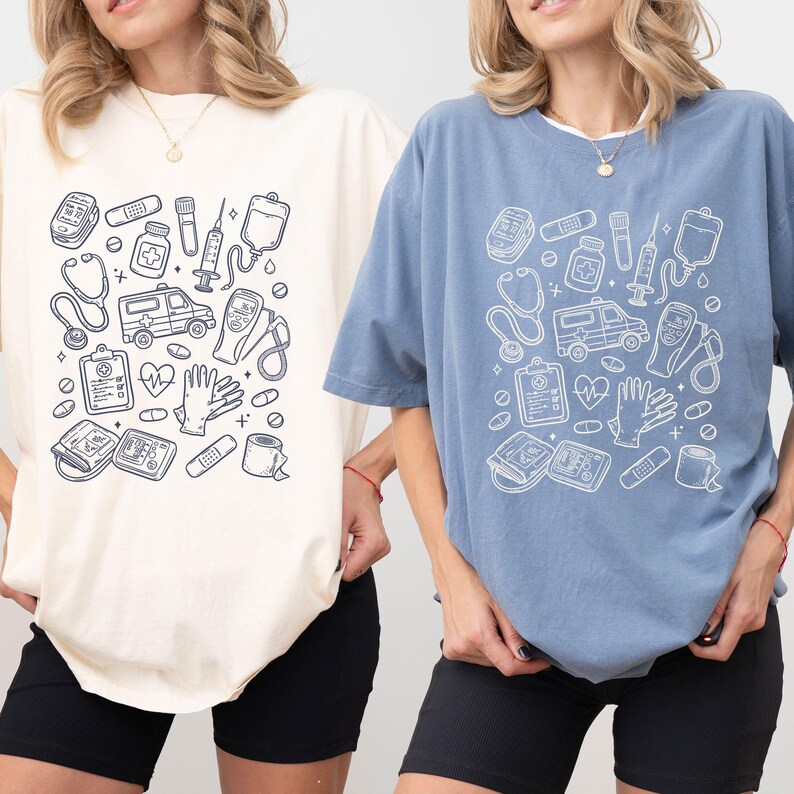 May include: Two t-shirts, one cream and one blue, featuring a white medical-themed graphic. The design includes a stethoscope, ambulance, syringe, and other medical equipment. The shirts are short-sleeved and have a relaxed fit.