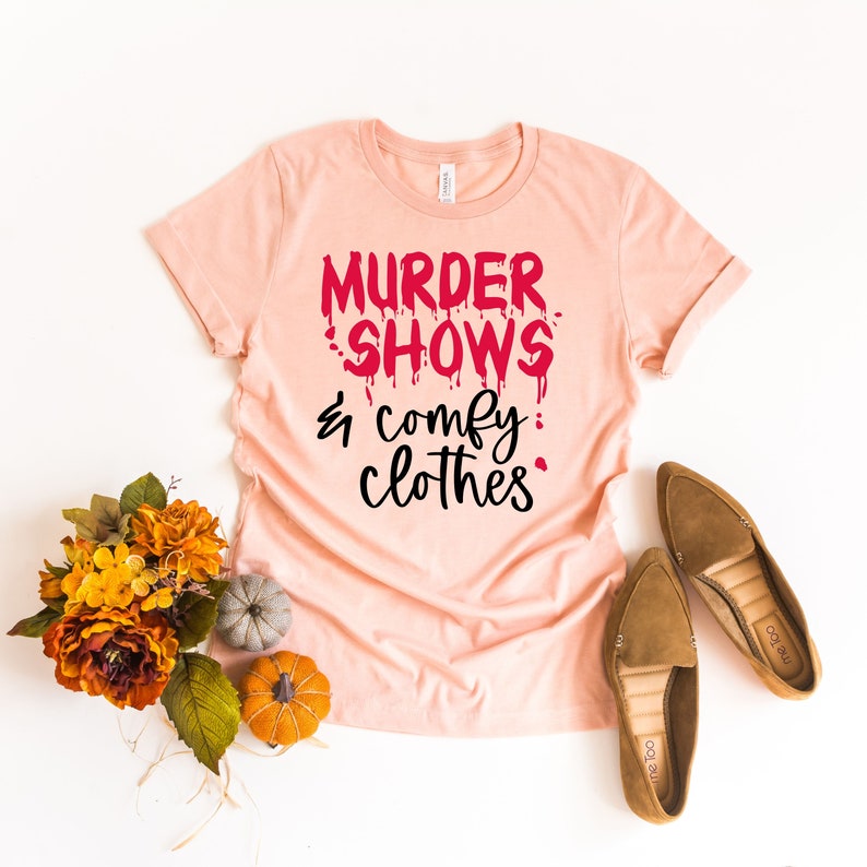 May include: A peach-colored t-shirt with the text "MURDER SHOWS & comfy clothes" in red and black. The shirt is styled with brown loafers, decorative pumpkins, and a fall floral arrangement.