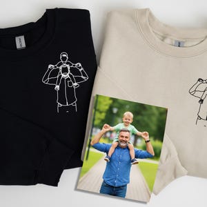 May include: Black and beige crewneck sweatshirts with a line drawing of a person carrying a child on their shoulders. A framed photo shows a man carrying a child on his shoulders. The sweatshirts are made of a soft material.