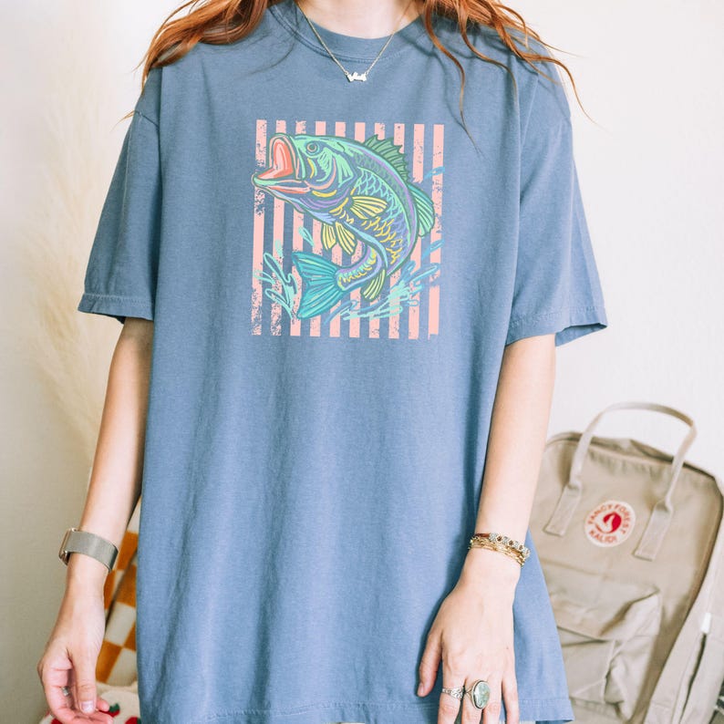 Pu&ograve; includere: T-shirt blu a maniche corte con una grafica colorata di un pesce che salta fuori dall'acqua. Il pesce &egrave; in tonalit&agrave; di blu, verde e giallo, su uno sfondo a strisce rosa e bianche. La maglietta ha una vestibilit&agrave; ampia.