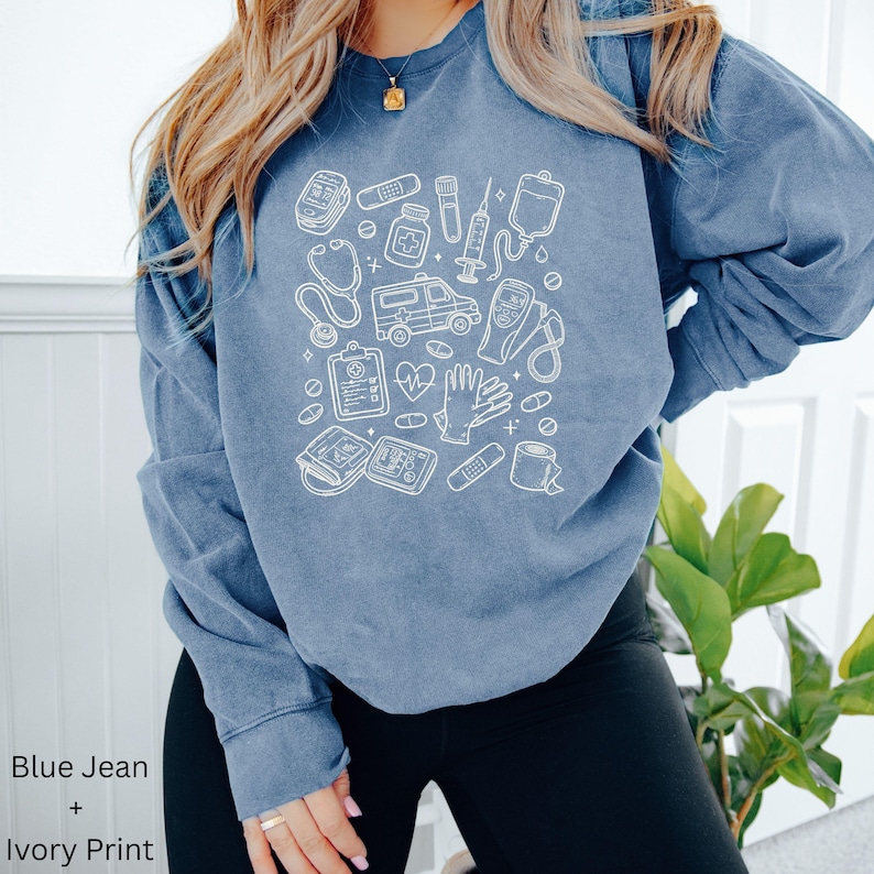 May include: Blue jean colored sweatshirt with an ivory print of medical equipment, including a stethoscope, ambulance, and medicine bottles. The sweatshirt has long sleeves and a crew neck. The text "Blue Jean + Ivory Print" is at the bottom left.