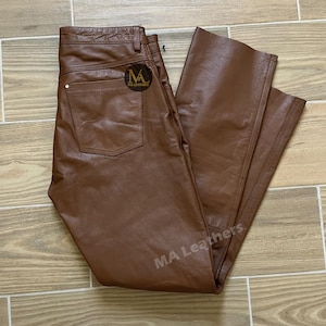 Puede incluir: Pantalones de cuero marrón con un diseño clásico de cinco bolsillos. Los pantalones tienen un corte de pierna recta y cierre de botón y cremallera. La marca "MA Leathers" es visible en el bolsillo trasero y la pierna.