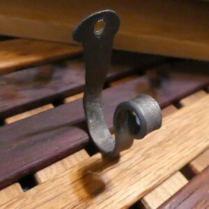May include: A rustic, dark metal hook with a curved design. The hook has a small, circular hole at the top for mounting. The hook is set against a wooden background with alternating dark and light brown slats.