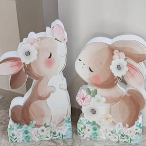 Könnte beinhalten: Zwei dekorative Hasenfiguren. Ein Hase hat einen Schmetterling auf dem Kopf, der andere hält Blumen. Beide Hasen sind braun und weiß mit floralen Akzenten und sitzen auf einer Basis mit floralem Design.