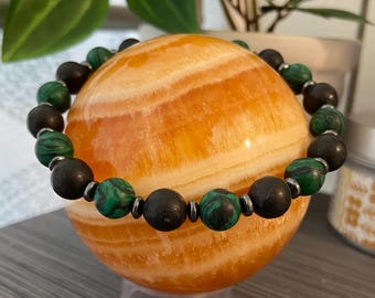 May include: A beaded bracelet with black and green malachite beads and dark brown beads. The bracelet is displayed on an orange sphere. The sphere is sitting on a clear stand.