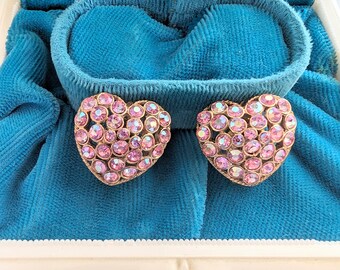 Op de afbeelding: Een paar hartvormige oorbellen met roze strass stenen gezet in verguld metaal. De oorbellen worden gepresenteerd in een blauwe fluwelen juwelendoos.