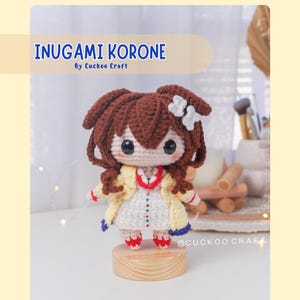 Puede incluir: Una muñeca amigurumi de crochet de una niña con cabello castaño, que lleva una chaqueta amarilla y un vestido blanco con detalles azules y rojos. La muñeca está de pie sobre una base de madera. El texto "INUGAMI KORONE by Cuckoo Craft" está en la parte superior de la imagen. El texto "@Cuckoo Craft" está en la parte inferior de la imagen.