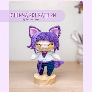 Può includere: Un personaggio all'uncinetto viola e bianco con un aspetto felino. Il personaggio indossa una camicia bianca, pantaloni viola e una coda viola. Il personaggio è in piedi su una base di legno. Il testo "CHENYA PDF PATTERN By Cuckoo Craft" è visualizzato nella parte superiore dell'immagine.