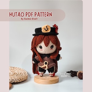 Puede incluir: Patrón de crochet para una muñeca amigurumi Hu Tao. La muñeca lleva un atuendo marrón y negro con un sombrero rojo y un ancla dorada. La muñeca está de pie sobre una base de madera. El texto "Hu Tao PDF Pattern" y "By Cuckoo Craft" es visible en la parte superior de la imagen.