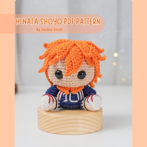 May include: Crochet pattern for a Hinata Shoyo amigurumi doll. The doll is wearing a blue and white jersey with orange hair. The doll is sitting on a wooden base. The pattern is available as a PDF download.