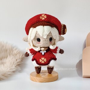 May include: A crocheted doll with white hair, a red hat with a brown trim and a brown flower, and a red dress with brown flowers. The doll is standing on a wooden base. The doll is wearing brown boots. The text "@cuckoocraft" is visible in the image.