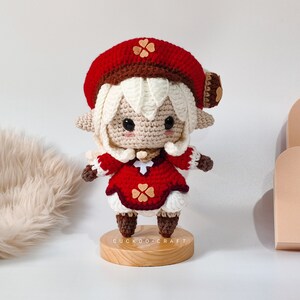 May include: A crocheted doll with white hair, a red hat with brown trim, and a red dress with brown trim. The doll is standing on a wooden base with the text "Cuckoo Craft".