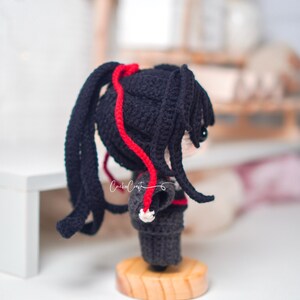 May include: A crocheted doll with black pigtails, detailed with red yarn. The doll is dressed in a dark outfit and stands on a wooden base. The doll's face is visible, with a small eye. The text "Crochet Craft" is visible.