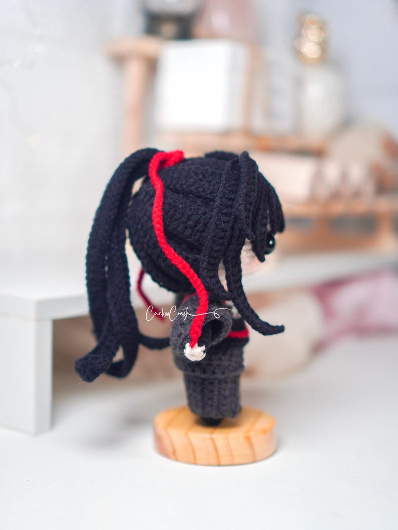 May include: A crocheted doll with black pigtails, detailed with red yarn. The doll is dressed in a dark outfit and stands on a wooden base. The doll's face is visible, with a small eye. The text "Crochet Craft" is visible.