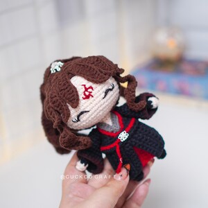 Op de afbeelding: Handgemaakte gehaakte pop met bruin haar en een zwart-rode outfit. De pop heeft een cr&egrave;mekleurig gezicht met geborduurde kenmerken en een decoratieve haarversiering. Een uniek, handgemaakt item.