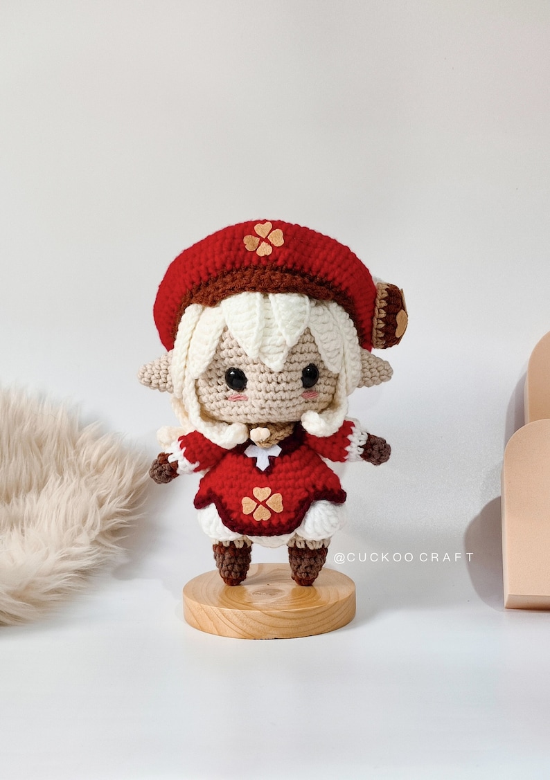 May include: A crocheted doll with white hair, a red hat with a brown trim and a brown flower, and a red dress with brown flowers. The doll is standing on a wooden base. The doll is wearing brown boots. The text "@cuckoocraft" is visible in the image.