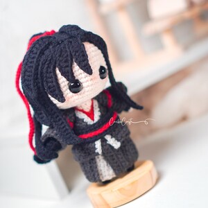 May include: A handcrafted crochet doll with black hair and red detailing. The doll is dressed in a dark grey outfit with red trim and a white under-layer. It stands on a small, circular wooden base. The doll has a charming, cartoon-like aesthetic.