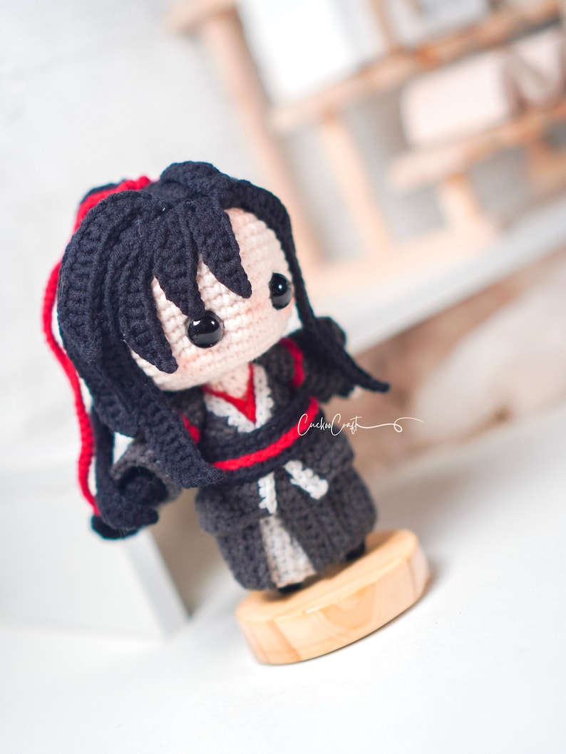 May include: A handcrafted crochet doll with black hair and red detailing. The doll is dressed in a dark grey outfit with red trim and a white under-layer. It stands on a small, circular wooden base. The doll has a charming, cartoon-like aesthetic.