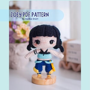 Puede incluir: Una muñeca de ganchillo con cabello negro y un atuendo azul y amarillo se encuentra sobre una base de madera. La muñeca tiene ojos grandes y está etiquetada como "ZOEY PDF PATTERN" por Cuckoo Craft.