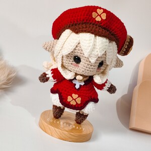 May include: Crocheted doll with white hair, a red beret with brown trim, a red dress with brown trim and white accents, and brown boots. The doll is standing on a wooden base with the text "Cuckoo-Craft" printed on it.