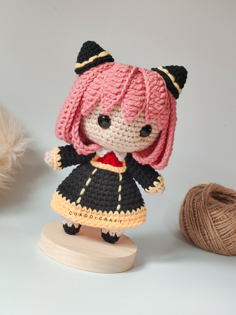 K&ouml;nnte beinhalten: Eine geh&auml;kelte Puppe mit rosa Haaren, schwarzen und gelben Kleidern und schwarzen Ohren. Die Puppe steht auf einem Holzsockel. Die Puppe ist handgefertigt und hat einen s&uuml;&szlig;en, fantasievollen Ausdruck.