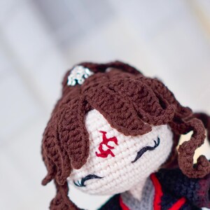 Op de afbeelding: Handgemaakte gehaakte pop met bruin haar en een zwarte outfit. De pop heeft een wit gezicht met rode geborduurde markeringen en zwarte wimpers. Het haar van de pop is gestyled in krullen. De tekst "@CUCKOO CRAFT" is zichtbaar.