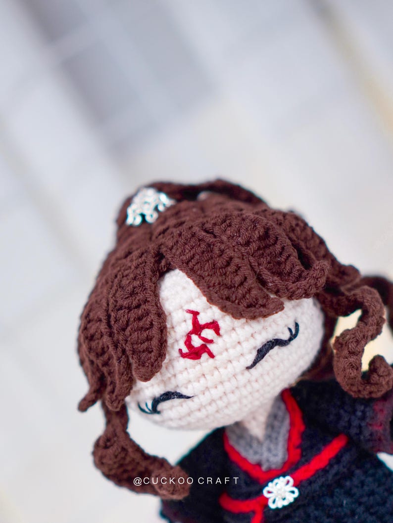 Op de afbeelding: Handgemaakte gehaakte pop met bruin haar en een zwarte outfit. De pop heeft een wit gezicht met rode geborduurde markeringen en zwarte wimpers. Het haar van de pop is gestyled in krullen. De tekst "@CUCKOO CRAFT" is zichtbaar.