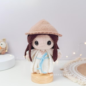 May include: A crocheted doll wearing a white dress and a brown conical hat. The doll has brown hair and large brown eyes. The doll is standing on a wooden base. The doll is a handmade craft item.