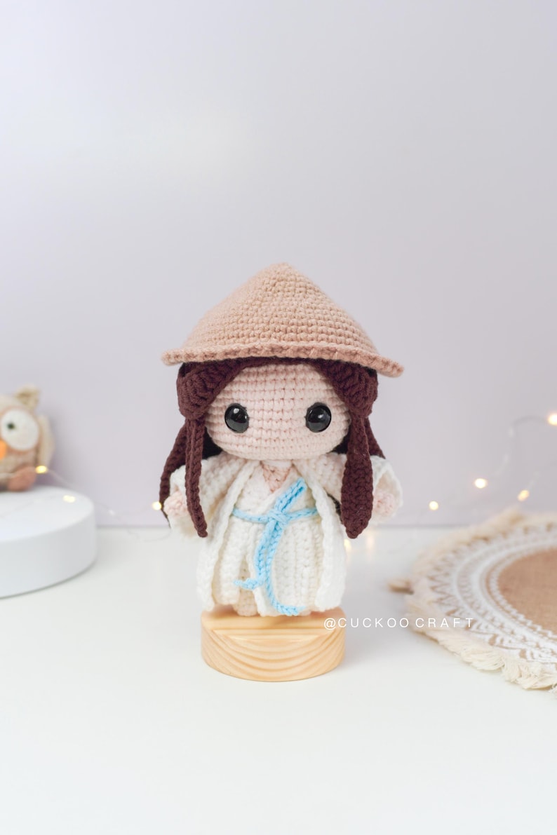 May include: A crocheted doll wearing a white dress and a brown conical hat. The doll has brown hair and large brown eyes. The doll is standing on a wooden base. The doll is a handmade craft item.