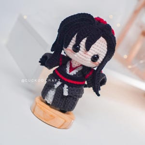 May include: A handcrafted crochet doll with black hair and a red detail. The doll is dressed in a black and grey outfit with red trim, mounted on a wooden base. The doll's face is pale pink with black button eyes. The text "@CUCKOO CRAFT" is visible.
