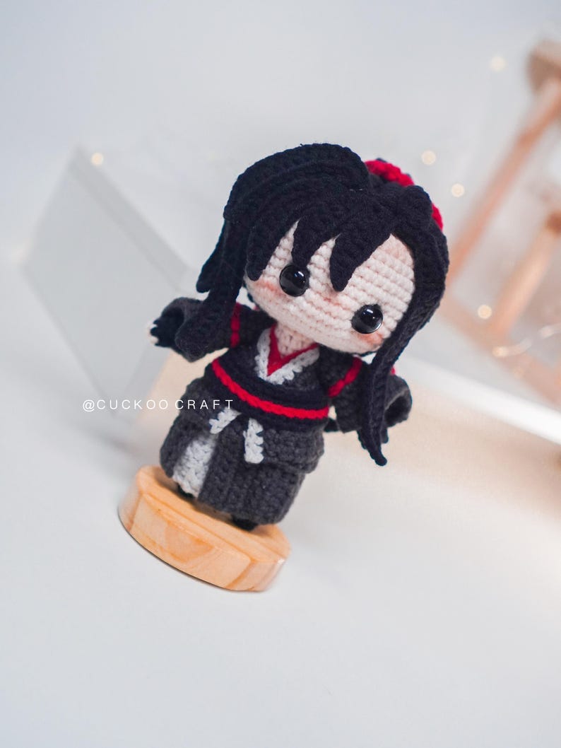 May include: A handcrafted crochet doll with black hair and a red detail. The doll is dressed in a black and grey outfit with red trim, mounted on a wooden base. The doll's face is pale pink with black button eyes. The text "@CUCKOO CRAFT" is visible.
