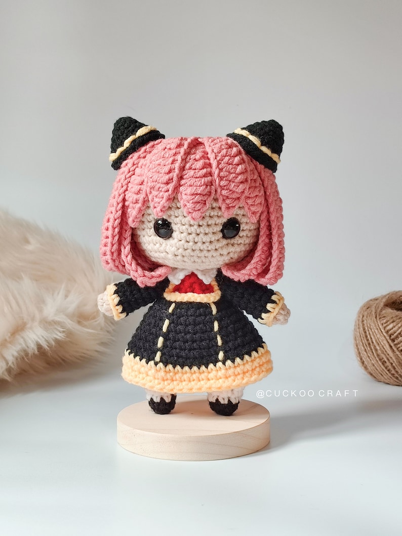 K&ouml;nnte beinhalten: Geh&auml;kelte Amigurumi-Puppe mit rosa Haaren, schwarzen Ohren und einem schwarzen und gelben Kleid. Die Puppe steht auf einem Holzsockel.
