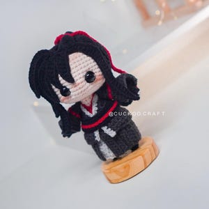 May include: A handmade crochet doll with black hair and a red accent, wearing a black and grey outfit. The doll is standing on a wooden base. The text "@CUCKOO CRAFT" is visible.