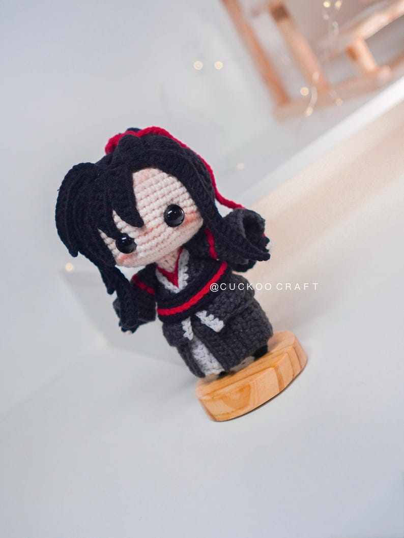 May include: A handmade crochet doll with black hair and a red accent, wearing a black and grey outfit. The doll is standing on a wooden base. The text "@CUCKOO CRAFT" is visible.